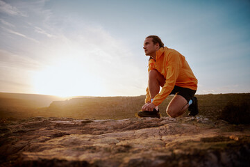 Fit male jogger tying shoelace preparing for cross country mountain trail in nature and looking away against sunrise © StratfordProductions