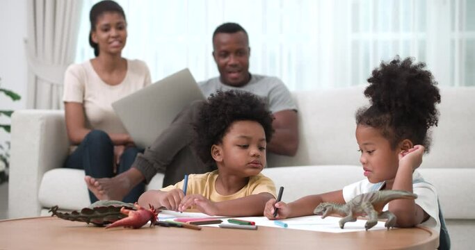 African American dad and mom sitting on sofa with computer and looking at their kids drawing a picture of dinosaurs with a smile in living room. Family and Development for kids concept.