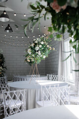 A large, tall arrangement of artificial flowers in white and peach on an empty table with a blue tablecloth. White room, sunny day.