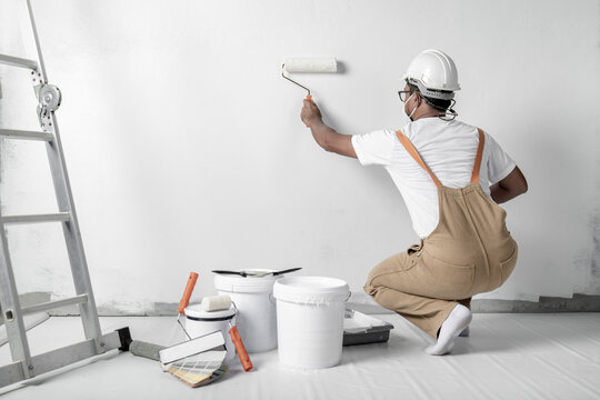 A Man Paints A White Wall With A Roller. Repair Of The Interior. Young Male Decorator Painting A Wall In The Empty Room, Concept Builder Or Painter In Helmet With Paint Roller Over The Empty Room