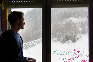 joven blanco y apuesto mirando por la ventana nevada