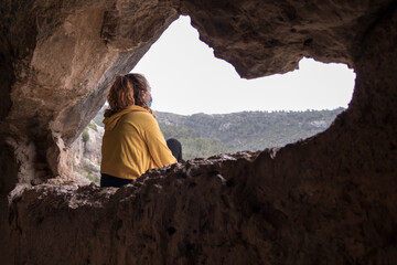Woman from behind with a yellow jacket sitting in a cave enjoying the scenery