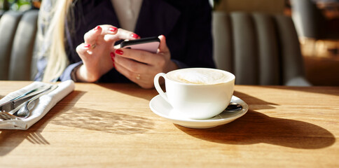 Woman typing text message on smart phone in a cafe. Young woman sitting at a table with a coffee using mobile phone. Coffee break. Cup of coffee on the table. Blurred image, selective focus