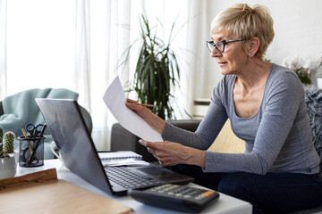 An older woman with short blonde hair sits on sofa in living room and works on laptop