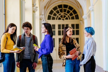 Fototapeta premium Cheerful diverse students discussing information from university standing indoors and talking, youth generation Z reading and smiling indoor at hall.