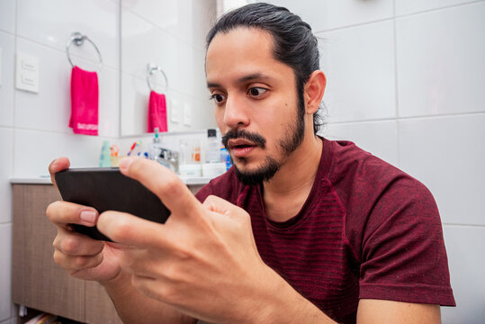 Young latin man smiling on the toilet using a smart phone at the bathroom. Man using the smartphone to play video games at the bathroom sitting on the toilet.