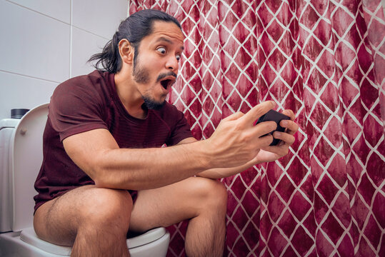 Young latin man smiling on the toilet using a smart phone at the bathroom. Man using the smartphone to play video games at the bathroom sitting on the toilet.
