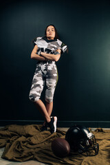 Athletic woman poses in American football uniform near helmet and ball.