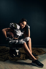 Athletic woman sits on helmet in American football uniform and holds ball.