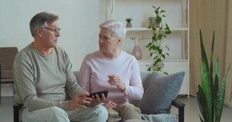 Elderly couple old wife woman and husband looking at mobile phone sitting on couch at home in living room arguing about what they saw on video online discussing received SMS message evil quarrel