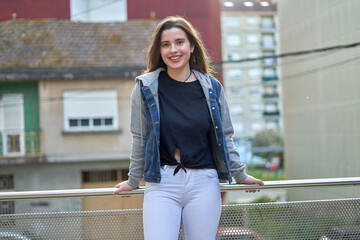 Young girl with long hair and light skin wearing a blue denim jacket leaning on a railing looking at the camera with unfocused urban background. teenager with street look in a city