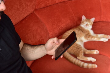 Boy sitting on red sofa using cell phone with cat sleeping beside.