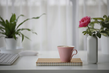 Pink coffee cup and red rose with notebook and plant pot on white wooden table
