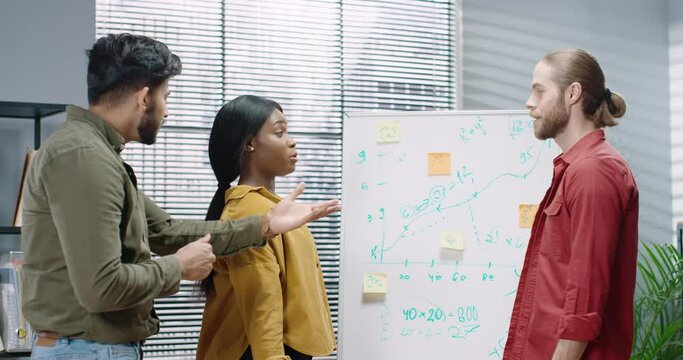 Multinational group of office workers are arguing while working on project infographic near mudboard. African American girl yells at guy in loudspeaker.