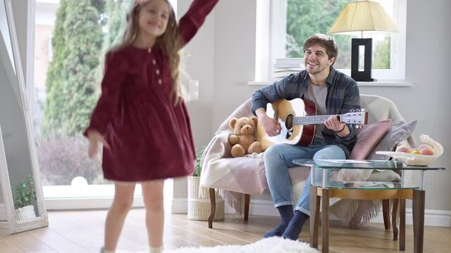 Wide shot of happy young brunette man playing guitar for cute little girl dancing at home. Cheerful pretty Caucasian daughter hugging loving father. Bonding and family concept