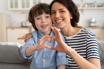 Mom and son making heart hand gesture together happy smiling look in camera at home sitting on...