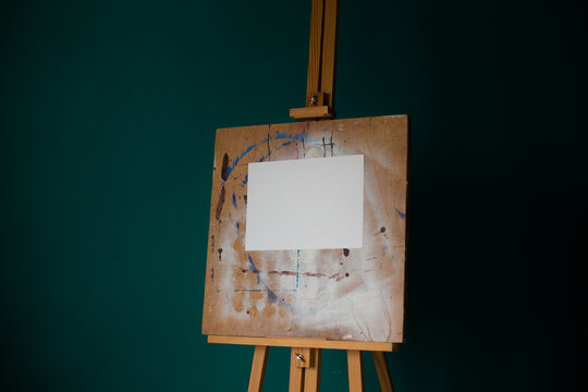Mock up of a white sheet of watercolor paper on an easel against a dark green wall background.
