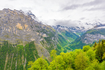 Naklejka premium View of beautiful landscape in the Alps with fresh green meadows and snow-capped mountain tops in the background on clouds in springtime.
