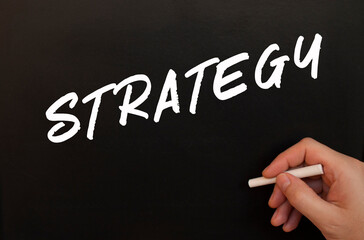 Male hand writes in chalk the words STRATEGY on a black board.