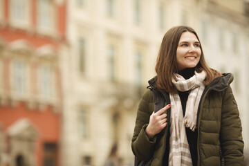 Travel concept: happy smiling young woman posing at street of European city. Model wearing green coat, white and beige scarf. Copy, empty space for text. Outdoor shot