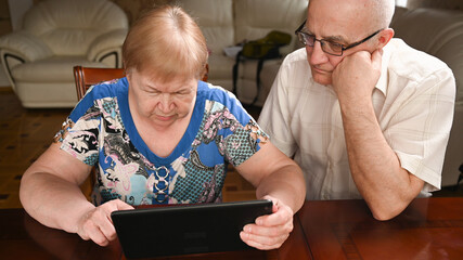 An elderly couple is sitting at a tablet and looking for information on the Internet.