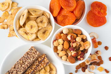 Banana chips, dried apricots, various nuts and granola bar in white plates on a light background.