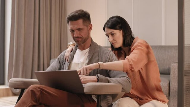 Medium long of Caucasian man sitting in armchair, typing on laptop computer, young dark-haired woman hugging him, pointing finger on screen, couple smiling