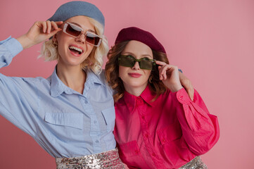 Two happy smiling girls wearing stylish sunglasses, colorful clothes posing on pink background. Copy, empty space for text