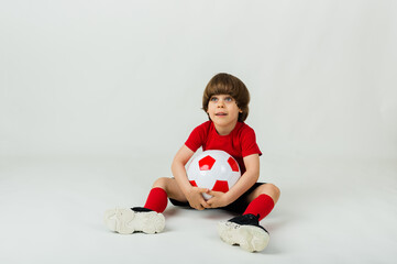 joyful little boy in uniform sits with a soccer ball on a white background with space for text