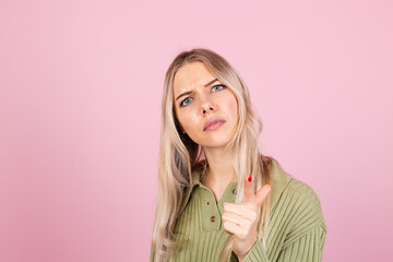 Pretty european woman in casual sweater on pink background angrily, reproachfully accuses you, points directly to camera, blames you