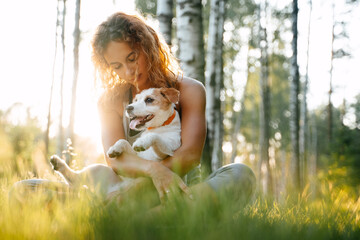 Young red-haired woman sitting on green grass in the park holding jack russell dog in her arms.