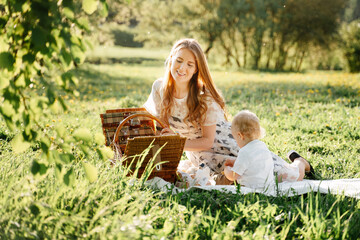 Happy mom with little daughter sits on a green lawn in the park on a picnic with baskets. Family day of parents with children.