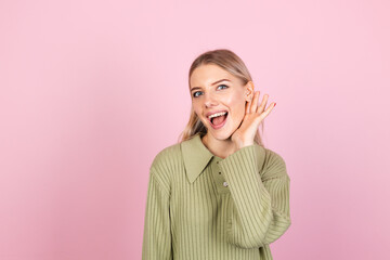 Pretty european woman in casual sweater on pink background curious try to hear what you saying with hand by ear copy space