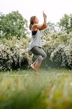 Full length side view of an athletic woman standing on one leg practicing yoga pose in a green park.