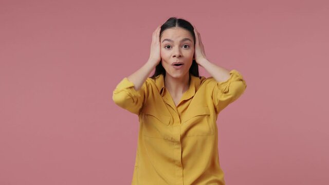 Excited shocked young latin woman 20s in yellow shirt posing isolated on pastel pink background studio. People lifestyle concept. Put hands on head cheeks say wow doing winner gesture spreading hands
