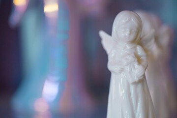 Fototapeta premium Small white marble figurine of an angel on a pearlescent pink blue blurred bokeh background. Place for text