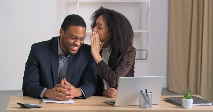 Afro american colleagues man and woman sitting in modern office in business company girl tells secret to male colleague share funny gossip joke good news confidential information man laughs rejoices