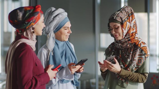 Three cheerful muslim businesswomen in hijabs standing in office and discussing something on smartphone screen