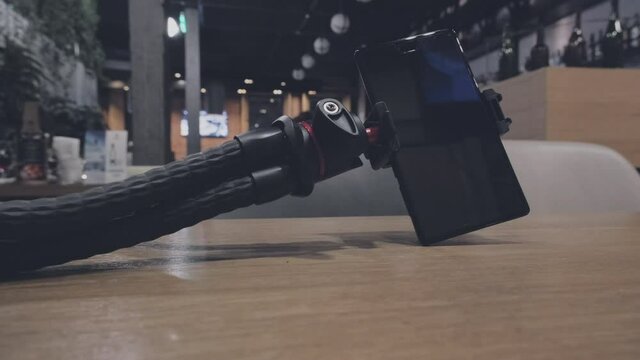 On the table in the restaurant is lies a phone on a tripod. A man hand picks up a tripod. The concept of a blogger restaurant critic and reviews of food establishments