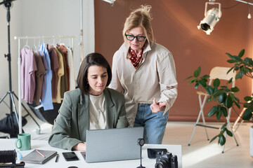 Young designer sitting at the table with laptop and discussing online work together with her colleague at studio