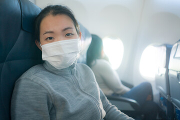 Fototapeta na wymiar Asian tourist woman flying in times of covid19 - young happy and excited Korean girl in face mask sitting on airplane cabin ready for flight looking at the camera