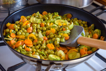 Process of cooking saute at home. Stewed vegetables and silicone spatula with wooden handle in frying pan. Mixed Ingredients leek, carrot, zucchini, bell pepper, green peas. Close-up. Selective focus.