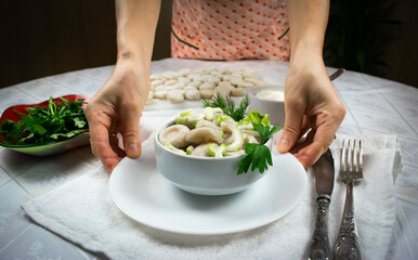 Hands of a woman in an apron hold out a bowl of cooked homemade dumplings, decorated with green leaves of dill and parsley. Selective focus.