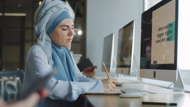 Side view of young muslim woman in hijab watching business presentation on computer and taking notes while working in office