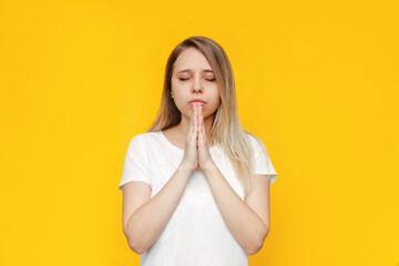 A young caucasian beautiful blonde woman in a white t-shirt prays with closed eyes and folded hands, thanks, making wish, asking help, hope or forgiveness isolated on a bright color yellow background