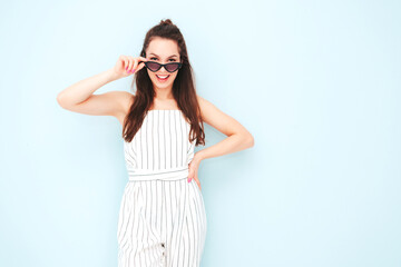 Portrait of young beautiful smiling female in trendy summer hipster overalls clothes. Sexy carefree woman posing near light blue wall in studio. Positive model having fun indoors.Taking off sunglasses