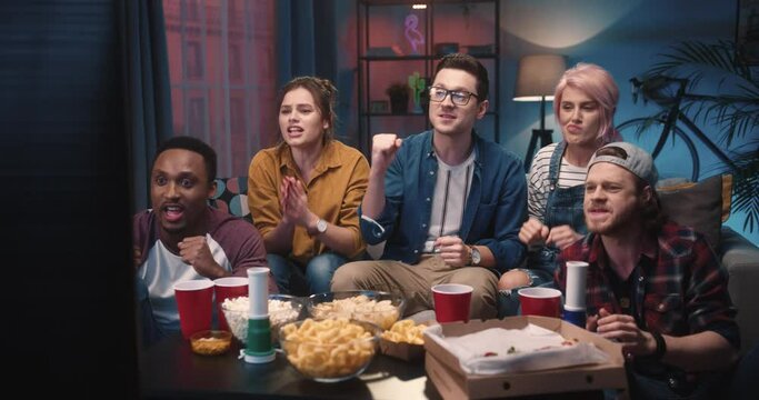 Joyful young mixed-race male and female friends watching sport match on TV at home in evening holding ball in hands feeling excited emotionally supporting soccer team and shouting when it score goal
