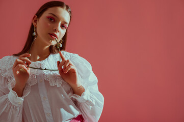 Fashionable elegant girl wearing trendy white vintage cotton blouse with lace collar, stylish pearl earrings, holding glasses, posing on pink background. Copy, empty space for text