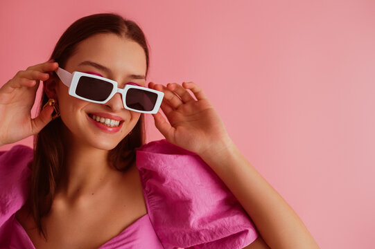 Fashionable girl wearing trendy rectangular sunglasses, pink blouse, posing on pink background. Close up studio portrait. Copy, empty space for text