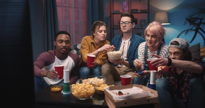 Mixed-race young cheerful company sitting on couch in living room gathered together drinking and eating snacks while watching movie on TV. Friends watching sport game in house. Entertainment concept